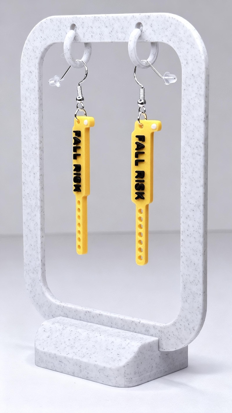 May include: Yellow earrings with the text "FALL RISK" in black lettering. The earrings are rectangular with a series of small holes along the bottom edge. They hang from silver-coloured ear wires and are displayed on a white stand.