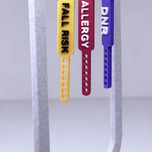 May include: Three medical alert earrings with the words "FALL RISK", "ALLERGY", and "DNR" printed on them. The earrings are yellow, red, and purple. Each earring has a silver hook.