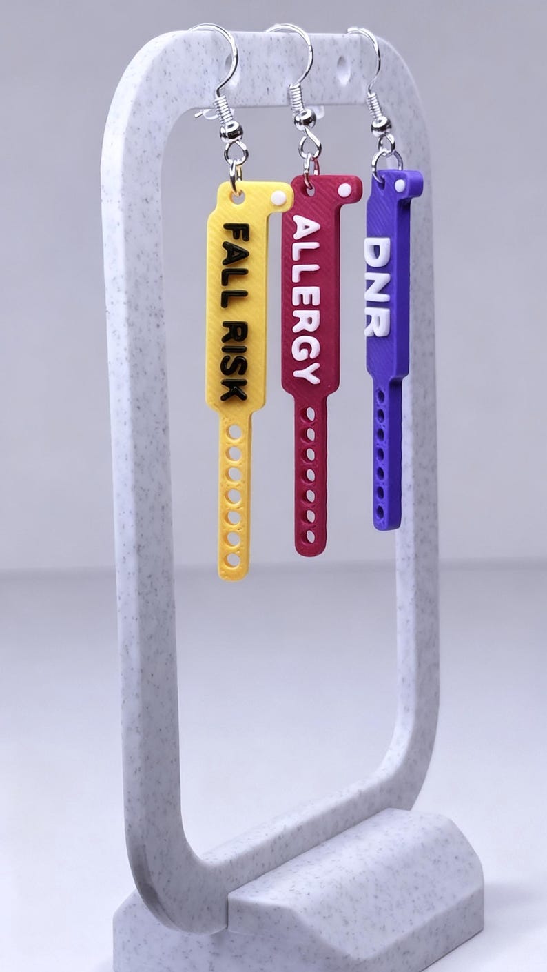 May include: Three medical alert earrings with the words "FALL RISK", "ALLERGY", and "DNR" printed on them. The earrings are yellow, red, and purple. Each earring has a silver hook.