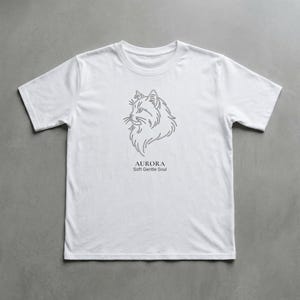 Può includere: T-shirt bianca con un disegno a tratto grigio della testa di un gatto. Sotto l'immagine c'è la parola "AURORA" e il testo "Soft Gentle Soul". La maglietta ha maniche corte ed è realizzata in un materiale morbido.
