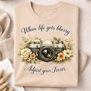 May include: A beige sweatshirt featuring a vintage camera design with floral accents in cream, peach, and green. The text "When life gets blurry, Adjust your Focus" is written above and below the camera illustration.