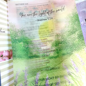 May include: Open Bible with a watercolor overlay featuring the text "You are the light of the world." The overlay depicts a field of lavender and green foliage. The Bible pages are open to Matthew 5:14.