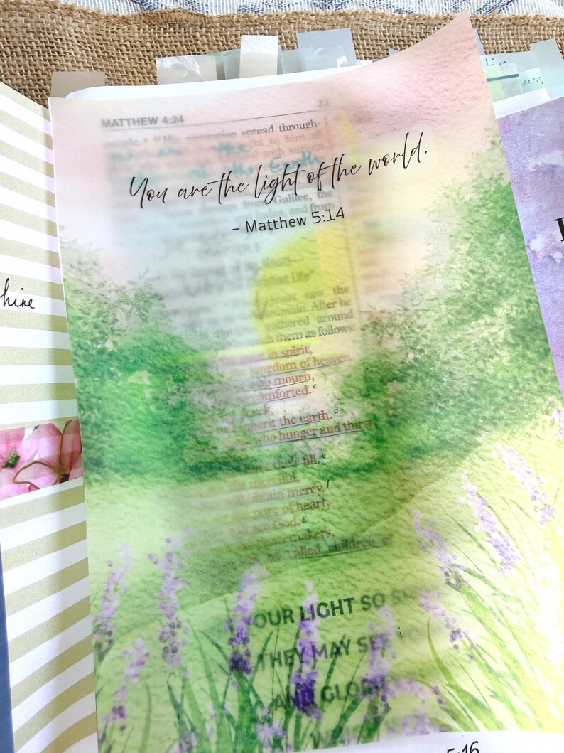 May include: Open Bible with a watercolor overlay featuring the text "You are the light of the world." The overlay depicts a field of lavender and green foliage. The Bible pages are open to Matthew 5:14.