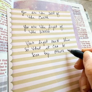 May include: A rectangular card with horizontal beige and white stripes and a scalloped left edge. The card features handwritten black ink text that reads: You are the salt of the Earth, You are the light of the World, Your light must shine so that it can be seen by others.