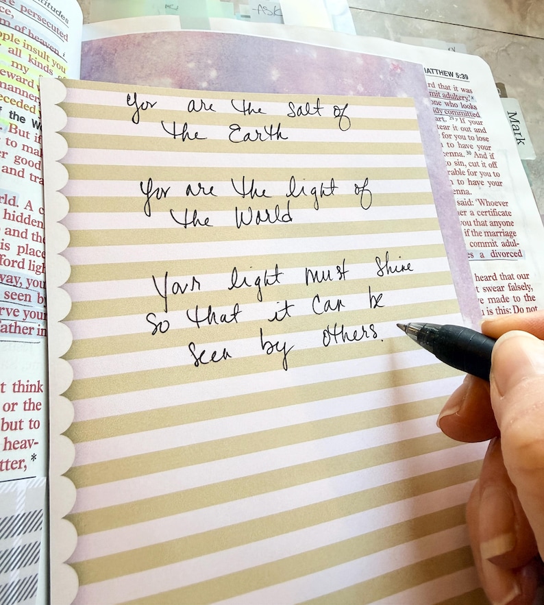 May include: A rectangular card with horizontal beige and white stripes and a scalloped left edge. The card features handwritten black ink text that reads: You are the salt of the Earth, You are the light of the World, Your light must shine so that it can be seen by others.