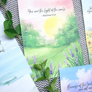 May include: A rectangular art print featuring a watercolor landscape of a sunlit green meadow with purple lavender flowers. The text reads: You are the light of the world. - Matthew 5:14. The scene includes a soft yellow sun and pink sky.