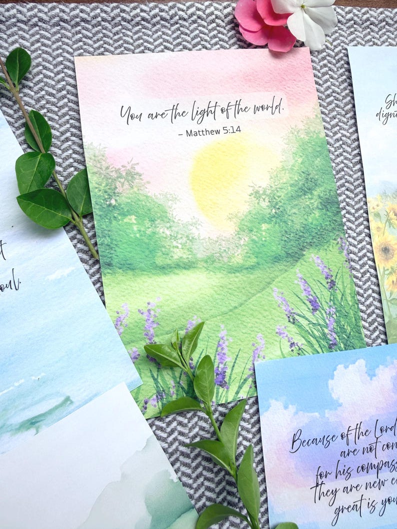 May include: A rectangular art print featuring a watercolor landscape of a sunlit green meadow with purple lavender flowers. The text reads: You are the light of the world. - Matthew 5:14. The scene includes a soft yellow sun and pink sky.
