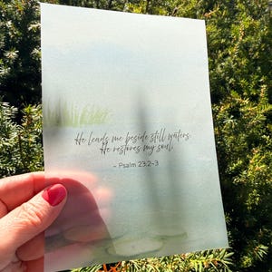 May include: A watercolor print with a serene landscape in shades of blue and green. The print features the text "He leads me beside still waters. He restores my soul. - Psalm 23:2-3" in elegant script. The print is held in front of a green bush.