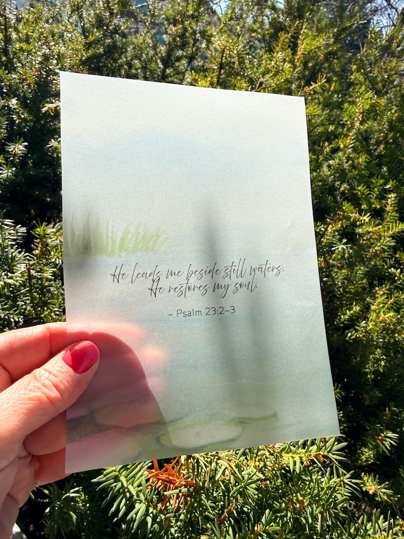 May include: A watercolor print with a serene landscape in shades of blue and green. The print features the text "He leads me beside still waters. He restores my soul. - Psalm 23:2-3" in elegant script. The print is held in front of a green bush.