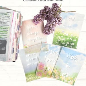 May include: A collection of watercolor floral Bible tip-ins with cut-out designs. The tip-ins feature phrases like "Grace changes everything" and "Held in His peace." A sprig of purple flowers rests on top of the tip-ins. The text "Quiet Grace Collection" is at the top.