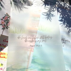 May include: Watercolor-style art print with the text "He leads me beside still waters. He restores my soul." from Psalm 23:2-3. The print features a soft, blurred landscape with green and blue hues, evoking a sense of peace.