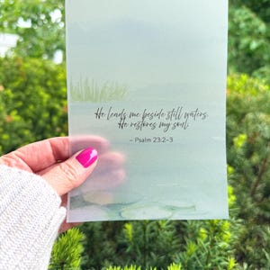 May include: A translucent card with a watercolor-style landscape and the text "He leads me beside still waters. He restores my soul. - Psalm 23:2-3." The card is held in front of a green, leafy background.