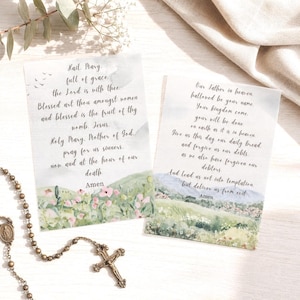May include: Two rectangular prayer cards featuring watercolor illustrations of rolling green hills and pink flowers. The left card displays the Hail Mary prayer, and the right card displays the Our Father prayer, both written in black cursive script.