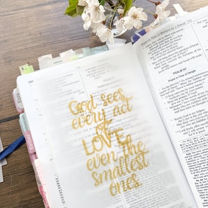 May include: An open Bible with a gold-colored overlay featuring the text "God sees every act, LOVE even the smallest ones." A small vase of white flowers sits in the background.