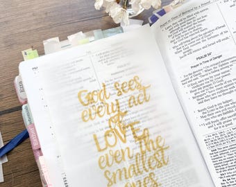 May include: An open Bible with a gold-colored overlay featuring the text "God sees every act, LOVE even the smallest ones." A small vase of white flowers sits in the background.