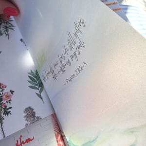 May include: Open book with watercolor illustrations and script. The text reads, "He leads me beside still waters. He restores my soul." Psalm 23:2-3 is also written. Floral and landscape art is visible.