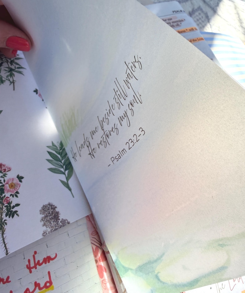 May include: Open book with watercolor illustrations and script. The text reads, "He leads me beside still waters. He restores my soul." Psalm 23:2-3 is also written. Floral and landscape art is visible.