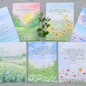 May include: A set of seven rectangular cards featuring watercolor illustrations of landscapes, flowers, and skies, each printed with a different Bible verse in cursive script. A small green leafy sprig is placed in the center of the arrangement.