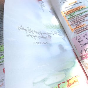 May include: Open Bible with handwritten notes and highlighted passages. The text includes Psalm 23 and other biblical verses. Colorful sticky notes and underlining highlight key phrases. The page is held open by a hand.