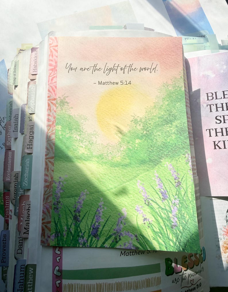 May include: A Bible page featuring a watercolor illustration of a sunlit green landscape with purple flowers. The text reads, "You are the light of the world. - Matthew 5:14." The page includes colorful index tabs along the side and additional text.