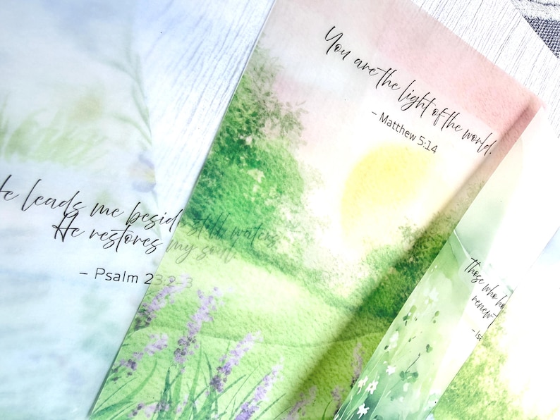 May include: Close-up of three watercolor-style bookmarks with inspirational quotes. One bookmark reads "You are the light of the world" from Matthew 5:14. Another says "He leads me beside still waters" from Psalm 23:2-3. The third bookmark has the text "Those who renew" from Isaiah.