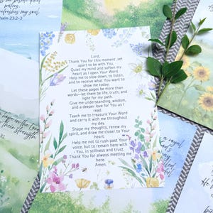 May include: A white rectangular card featuring a printed prayer in black text, surrounded by a colorful watercolor floral border with blue, yellow, pink, and green flowers. A small green leafy branch rests on the right side of the card.