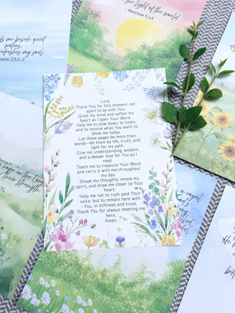 May include: A white rectangular card featuring a printed prayer in black text, surrounded by a colorful watercolor floral border with blue, yellow, pink, and green flowers. A small green leafy branch rests on the right side of the card.