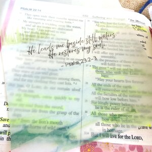 May include: Open Bible with highlighted text and a translucent overlay featuring the quote "He leads me beside still waters; He restores my soul" from Psalm 23:2-3. The page is marked with colorful tape.