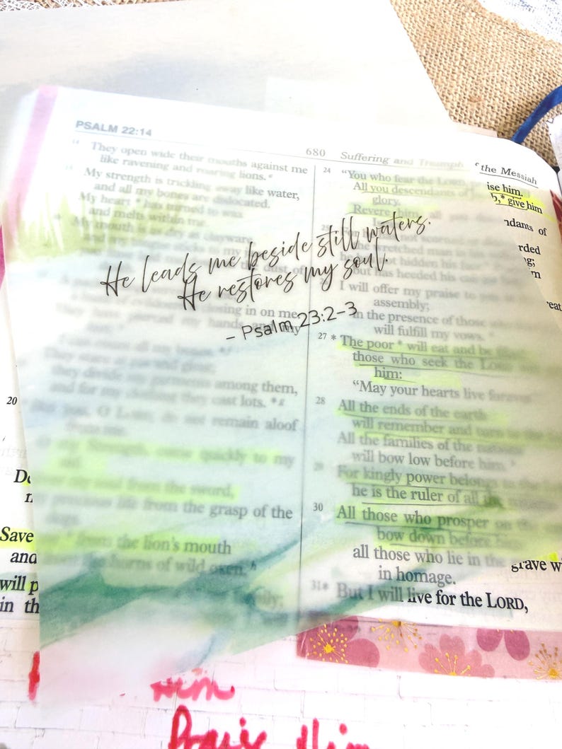 May include: Open Bible with highlighted text and a translucent overlay featuring the quote "He leads me beside still waters; He restores my soul" from Psalm 23:2-3. The page is marked with colorful tape.