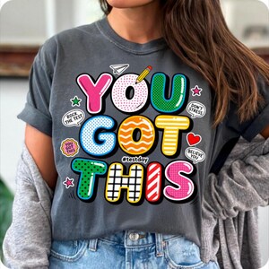 May include: Dark gray t-shirt with the words "YOU GOT THIS" in colorful, bubble-style letters. The design includes a pencil, paper airplane, stars, and encouraging phrases like "Rock the Test" and "Believe You".