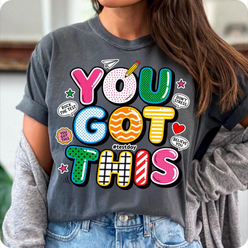 May include: Dark gray t-shirt with the words "YOU GOT THIS" in colorful, bubble-style letters. The design includes a pencil, paper airplane, stars, and encouraging phrases like "Rock the Test" and "Believe You".