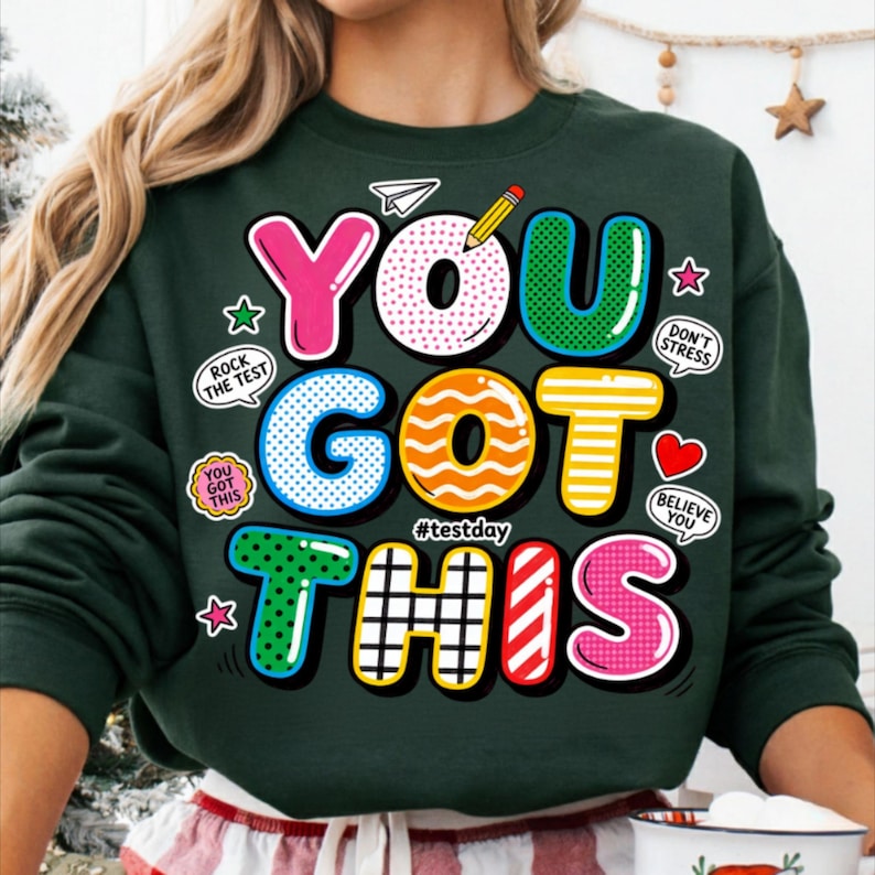 May include: Dark green sweatshirt with the words "YOU GOT THIS" in colorful, bubble-style letters. The design includes a pencil, paper airplane, stars, and encouraging phrases like "Rock the Test" and "Believe You". The hashtag #testday is also included.