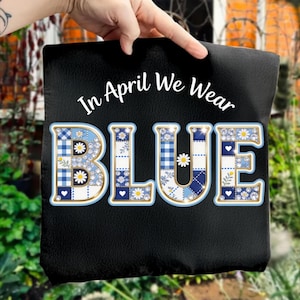以下が含まれることがあります： 「In April We Wear BLUE」の文字が入った黒いトートバッグ。「BLUE」の文字は大きく、青、白、金色のパッチワークデザインで、デイジーとハートが特徴です。