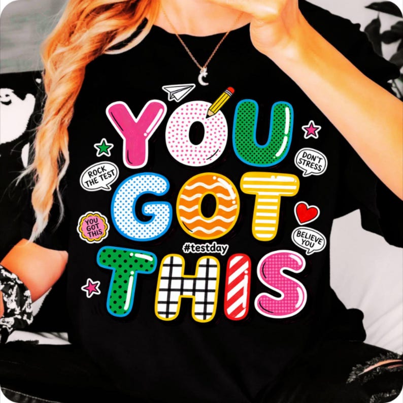 May include: Black t-shirt with a colorful graphic that says "YOU GOT THIS" in bubble letters. The design includes a pencil, paper airplane, stars, and the hashtag #testday. Additional text includes "Rock the Test", "You Got This", "Don't Stress", and "Believe You".