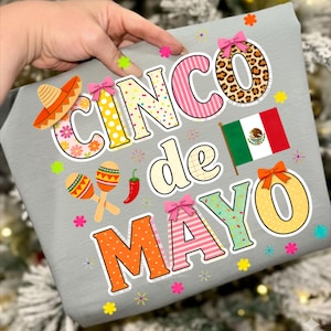 Può includere: Una maglietta grigia con la scritta "Cinco de Mayo" in lettere colorate e festive. Il design include un sombrero, maracas, un peperoncino e la bandiera messicana, con fiocchi decorativi e accenti floreali.