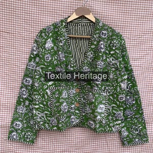 Könnte beinhalten: Eine gesteppte Jacke mit grün-weißem Blumenmuster, V-Ausschnitt, langen Ärmeln und zwei Vordertaschen. Die Jacke hat einen Knopfverschluss mit zwei sichtbaren Knöpfen und hängt an einem Kleiderbügel. Der Text Textile Heritage ist zu sehen.
