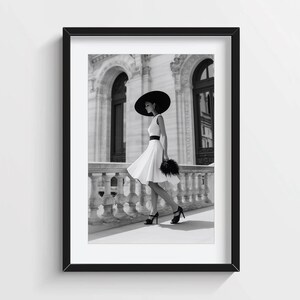 Può includere: Una stampa artistica incorniciata in bianco e nero che ritrae una persona con un cappello nero a tesa larga, un abito bianco senza maniche con fascia nera in vita e scarpe nere con tacco alto, su un balcone in pietra con colonne decorate.