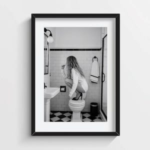 May include: A black and white photograph in a black frame showing a person with long hair crouching on a toilet in a bathroom. The room features white subway tile walls, a black and white chequered floor, a pedestal sink, and a hanging towel.
