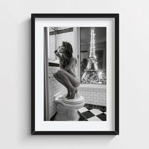 Può includere: Una stampa artistica incorniciata in bianco e nero che ritrae una persona intenta a truccarsi seduta su un WC in un bagno piastrellato. Dalla finestra è visibile la Torre Eiffel illuminata di notte, creando un contrasto con l'ambiente interno.