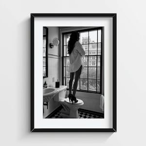 Può includere: Una fotografia incorniciata in bianco e nero che ritrae una persona in piedi su un water in un bagno, che indossa una camicia bianca abbottonata e scarpe nere con tacco, mentre guarda fuori da una grande finestra verso un edificio cittadino.