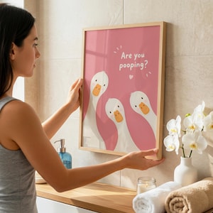 Op de afbeelding: Een ingelijste print met een roze achtergrond en de tekst "Are you pooping?" toont drie cartoon ganzen. De ganzen hebben witte veren en oranje snavels. Het frame is gemaakt van lichtgekleurd hout.