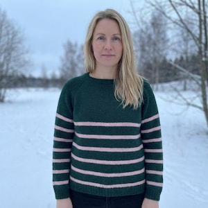May include: A green sweater with horizontal pink stripes. The sweater has a crew neck and long sleeves. The person is standing outside in a snowy field with trees in the background. The sweater is a knitwear item.