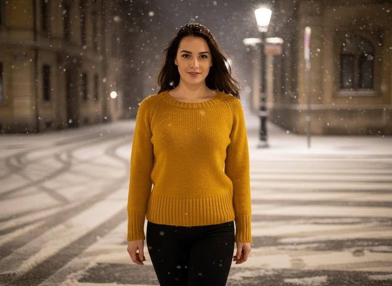 May include: A woman wearing a mustard yellow sweater and black pants stands on a snow-covered street at night. The sweater has a ribbed hem and cuffs. Snowflakes fall around her, and streetlights illuminate the scene.