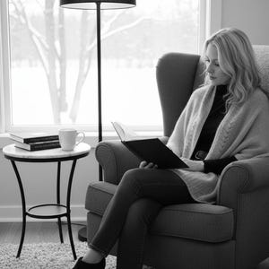 Puede incluir: Una persona sentada en un sillón gris, leyendo un libro. Lleva un suéter y ropa oscura. Una pequeña mesa auxiliar con tapa de mármol tiene libros y una taza. También se ven una lámpara de pie y una ventana con vistas nevadas.