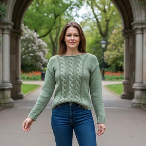 May include: A light green cable-knit sweater with long sleeves and a boat neck. The sweater features a central cable design and is paired with blue jeans. The background includes a stone archway and lush greenery.