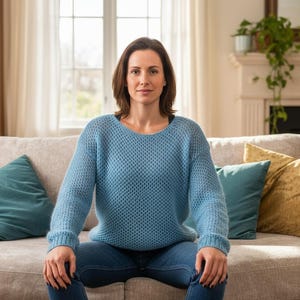 May include: A woman wearing a light blue knit sweater and blue jeans sits on a beige sofa. The sofa has teal and gold throw pillows. A bookshelf, window, and fireplace are visible in the background.