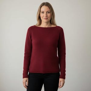 May include: A burgundy-colored sweater with a boat neck and long sleeves. The sweater has a ribbed texture and is paired with black pants. The woman is standing in front of a plain, light-colored background.