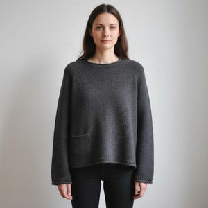 May include: A dark gray sweater with a crew neck and long sleeves. The sweater has a single pocket on the front and is made of a textured knit fabric. The model is wearing black pants.