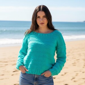 May include: A turquoise knit sweater with a round neckline. The long-sleeved sweater has a textured pattern and is worn with blue jeans. The background shows a beach with sand and ocean.