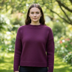May include: A plum-colored sweater with a ribbed neckline and cuffs. The long-sleeved knitwear has a classic design, suitable for cooler weather. The model is standing in a garden setting with a blurred background of green foliage and flowers.